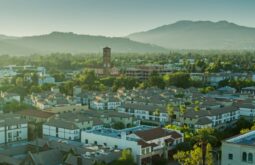 Aerial view of a suburban town with a tall brick church tower, surrounded by houses and palm trees, with mountains in the distance.