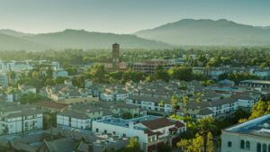Aerial view of a suburban town with a tall brick church tower, surrounded by houses and palm trees, with mountains in the distance.