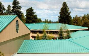 Low-angle view of a beige building with turquoise-green metal roofs and tall evergreen trees in the background.