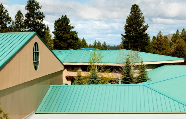 Low-angle view of a beige building with turquoise-green metal roofs and tall evergreen trees in the background.